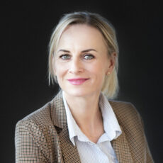 Katarzyna Zielant, z blond włosami związanymi z tyłu, uśmiecha się delikatnie w kraciastej marynarce na koszuli w paski. Ciemne tło idealnie podkreśla jej twarz.